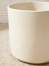 Load image into Gallery viewer, A close-up of a large, cylindrical off-white ceramic planter or container with a textured matte finish, resting on a woven beige rug.
