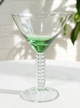 Load image into Gallery viewer, A clear glass martini-style cocktail glass with a ribbed stem and a green liquid inside, set on a white surface with soft leaf shadows and a blurred green plant in the background.
