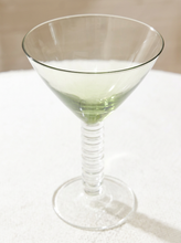 Load image into Gallery viewer, A clear glass martini coupe with a ribbed stem, shown on a white textured surface with a soft beige background.

