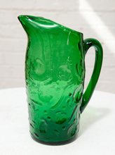 Load image into Gallery viewer, A vintage green glass pitcher with a textured, bubbly pattern and a curved handle, shown on a white surface against a white brick wall.
