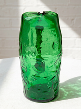 Load image into Gallery viewer, A translucent green glass vase with a textured, organic wavy surface, standing on a white fabric surface against a white brick wall with dappled sunlight.
