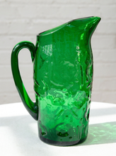 Load image into Gallery viewer, A vintage-style green glass pitcher with a textured, bubbled pattern and a curved handle, shown on a white surface against a white brick wall.
