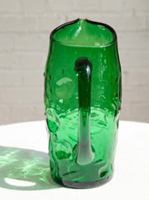 Load image into Gallery viewer, A green textured glass pitcher with a thick handle, sitting on a white surface with a white brick wall background.
