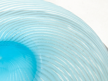 Load image into Gallery viewer, A close-up of a light blue ribbed glass plate or dish with concentric circular ridges and a brighter turquoise blue area in the lower left, set against a plain white background.
