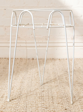 Load image into Gallery viewer, A white metal plant stand with a circular top frame and four tapered legs, shown on a woven jute rug against a white brick wall.

