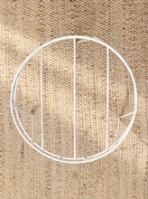 Load image into Gallery viewer, Overhead view of the white metal plant stand's circular wire frame and parallel support bars on a beige woven rug.
