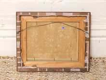 Load image into Gallery viewer, The back view of a framed canvas print, showing the wooden frame, backing board, hanging wire, and a small blue sticker, set against a white brick wall and woven rug.

