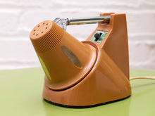 Load image into Gallery viewer, Orange desk lamp folded down into its base, showing the shade's vented back and retracted chrome stem on a green table.
