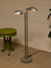 Load image into Gallery viewer, A modern two-light floor lamp with adjustable dome-shaped silver shades and a gray base, standing on a woven rug against a white brick wall next to a green side table.
