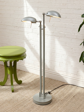Load image into Gallery viewer, A modern silver floor lamp with two adjustable dome-shaped shades on a round base, standing on a beige rug in front of a white brick wall next to a green side table.
