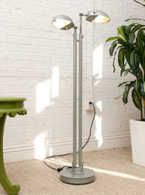 Load image into Gallery viewer, A modern silver floor lamp with two adjustable dome-shaped lights, standing on a beige carpeted floor against a white brick wall. A green plant in a white pot and a green side table are visible in the background.
