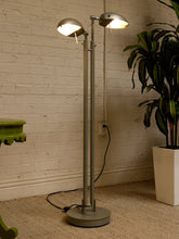 Load image into Gallery viewer, A modern two-light floor lamp with adjustable silver metal shades and a round base, shown lit in a room corner against a white brick wall with a potted plant nearby.
