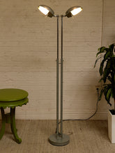 Load image into Gallery viewer, A tall floor lamp with two adjustable dome-shaped shades, shown lit in a room setting with a white brick wall, a green stool on the left, and a potted plant on the right.
