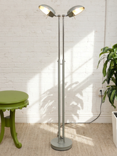 Load image into Gallery viewer, A tall dual-head floor lamp with a matte gray metal frame and silver dome-shaped shades, standing on a beige rug against a white brick wall. A green side table is on the left, and a potted plant is on the right.

