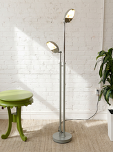 Load image into Gallery viewer, A tall dual-head floor lamp with adjustable silver metal arms and glowing warm white light heads, standing on a round base in a room with a white brick wall, beige rug, green stool, and potted plant.
