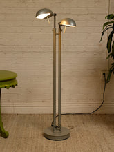 Load image into Gallery viewer, A tall industrial-style dual-head floor lamp with a circular base and two adjustable dome-shaped shades, shown in a room setting against a white brick wall with a green plant and stool partially visible.
