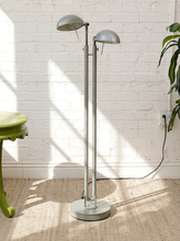 Load image into Gallery viewer, A modern two-light floor lamp with a brushed silver metal finish, standing on a round base against a white brick wall with sunlight casting shadows.
