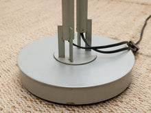 Load image into Gallery viewer, Close-up of a silver metal lamp base with vertical support rods and a black power cord, resting on a beige woven rug.
