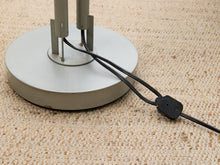 Load image into Gallery viewer, Close-up of a silver metal lamp base with black power cords running across a beige woven carpet.
