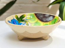 Load image into Gallery viewer, A handmade ceramic footed bowl with a cream-colored exterior and a colorful hand-painted interior featuring blue flowers, green leaves, and a dark brown crescent.
