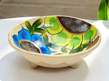 Load image into Gallery viewer, A small ceramic footed bowl with a hand-painted floral design inside, featuring blue flowers, green leaves, and a dark brown pattern on a cream-colored glaze, set on a white surface.
