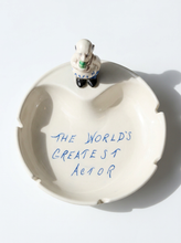 Load image into Gallery viewer, An off-white ceramic ashtray with the phrase 'THE WORLD'S GREATEST ACTOR' written in blue marker inside, topped with a small figurine of a bald man in a white suit and black shoes.
