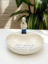 Load image into Gallery viewer, A white ceramic ashtray with a small figurine of a man holding a green bottle on top, with blue handwritten text reading 'THE WORLD'S CREEPEST' on the front, set on a white table with a green plant in the background.
