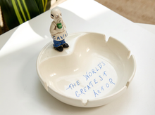 Load image into Gallery viewer, A cream-colored ceramic ashtray with a small figurine of a man holding a green drink on top, and blue handwritten text inside reading 'THE WORLD'S GREATEST APOOR'.
