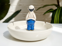 Load image into Gallery viewer, A ceramic figurine of a seated person with a white body, dark hair, and bright blue pants, positioned inside a white oval ceramic dish on a white surface with blurred green plants in the background.
