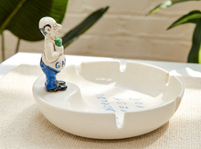 Load image into Gallery viewer, A white ceramic ashtray with a small figurine of a man holding a green apple on its hip, standing on the rim. The figurine has blue pants and black shoes, and the ashtray has blue writing inside.
