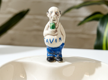 Load image into Gallery viewer, A small ceramic figurine of a bald man with blue pants holding a green object, standing on a glossy white surface. The figurine has the name 'SAVIN' written on the front.
