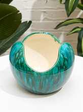 Load image into Gallery viewer, A ceramic planter bowl with a glossy teal and green marbled exterior and a cream-colored interior, displayed on a white surface with green plants and a white brick wall in the background.
