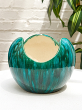Load image into Gallery viewer, A glossy ceramic bowl with a split rim and a marbled teal and green glaze, set on a white surface against a white brick wall with green plant leaves in the background.
