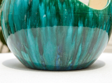 Load image into Gallery viewer, A close-up of a round ceramic bowl with a glossy, marbled teal and green glaze, resting on a white textured surface.
