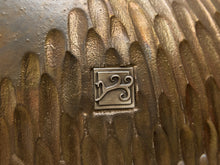 Load image into Gallery viewer, Close-up of a textured metallic surface with a small square metal emblem featuring a stylized symbol, likely part of a decorative object or hardware piece.
