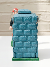 Load image into Gallery viewer, A small figurine of a person in a red helmet and black shirt peeking out from behind a blue brick-patterned box with a green base, photographed on a white surface against a light brick wall background.
