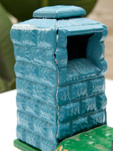 Load image into Gallery viewer, A small hand-painted blue trash can with a textured brick pattern, featuring a slanted opening and a matching lid, shown outdoors on a white surface with green foliage in the background.
