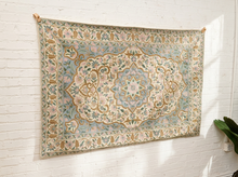 Load image into Gallery viewer, A large bohemian-style area rug with an intricate floral medallion pattern, hanging on a white brick wall.
