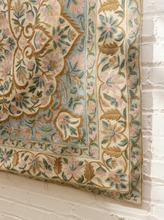 Load image into Gallery viewer, A close-up of an ornate area rug with a floral and medallion pattern in beige, dusty pink, light blue, and olive green, shown hanging against a white painted brick wall.
