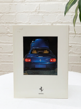 Load image into Gallery viewer, A square cream-colored picture frame featuring a photograph of a blue Ferrari sports car, displayed on a white surface against a white brick wall with a green plant leaf in the corner.
