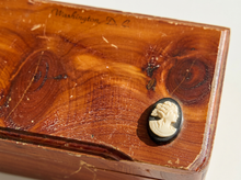 Load image into Gallery viewer, A close-up of a vintage wooden box lid with a carved cameo-style button or ornament on the corner, showing warm brown wood grain and faint handwritten text near the top.
