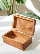 Load image into Gallery viewer, An open rectangular wooden box with a hinged lid, made from warm medium-brown wood with visible grain patterns, set on a white surface with soft natural shadows and blurred green leaves in the background.
