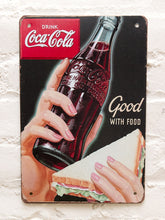 Load image into Gallery viewer, Vintage-style Coca-Cola advertising metal sign with black background, red Coca-Cola logo, illustrated hands holding a glass Coke bottle and sandwich, and Good With Food lettering

