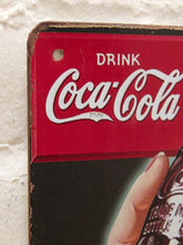 Load image into Gallery viewer, A close-up of a vintage-style Coca-Cola sign featuring the red and white Coca-Cola logo, mounted against a white textured wall. Part of a hand holding a Coca-Cola bottle is visible at the bottom right.
