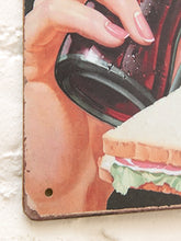 Load image into Gallery viewer, A close-up of a decorative metal sign featuring an illustration of a hand with pink manicured nails holding a glass of red wine next to a sandwich, mounted on a white textured wall.
