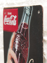Load image into Gallery viewer, A vintage-style Coca-Cola metal sign featuring a hand holding a glass bottle of cola, with the red Coca-Cola logo panel in the upper left, photographed against a white textured wall.
