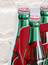 Load image into Gallery viewer, Vintage-style Coca-Cola metal advertising sign shaped like a six-pack of green glass Coke bottles in a red cardboard carrier, featuring white Coca-Cola lettering and retro soda graphics
