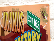 Load image into Gallery viewer, Rectangular Uranus Fudge Factory advertising metal sign with orange background, bold yellow and blue lettering, cartoon gentleman, green Exit 163 sign, and Historic Route 66 graphics
