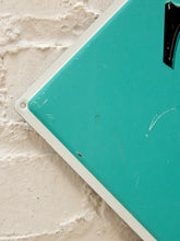 Load image into Gallery viewer, A close-up of a turquoise rectangular sign or board with a white border, placed against a white brick wall. A small black triangular piece is visible at the top right corner.
