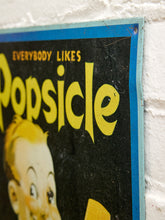 Load image into Gallery viewer, A close-up of a vintage-style poster or sign with the text 'EVERYBODY LIKES Popsicle' in yellow on a black background, showing part of a cartoon child's face, mounted against a white textured wall.
